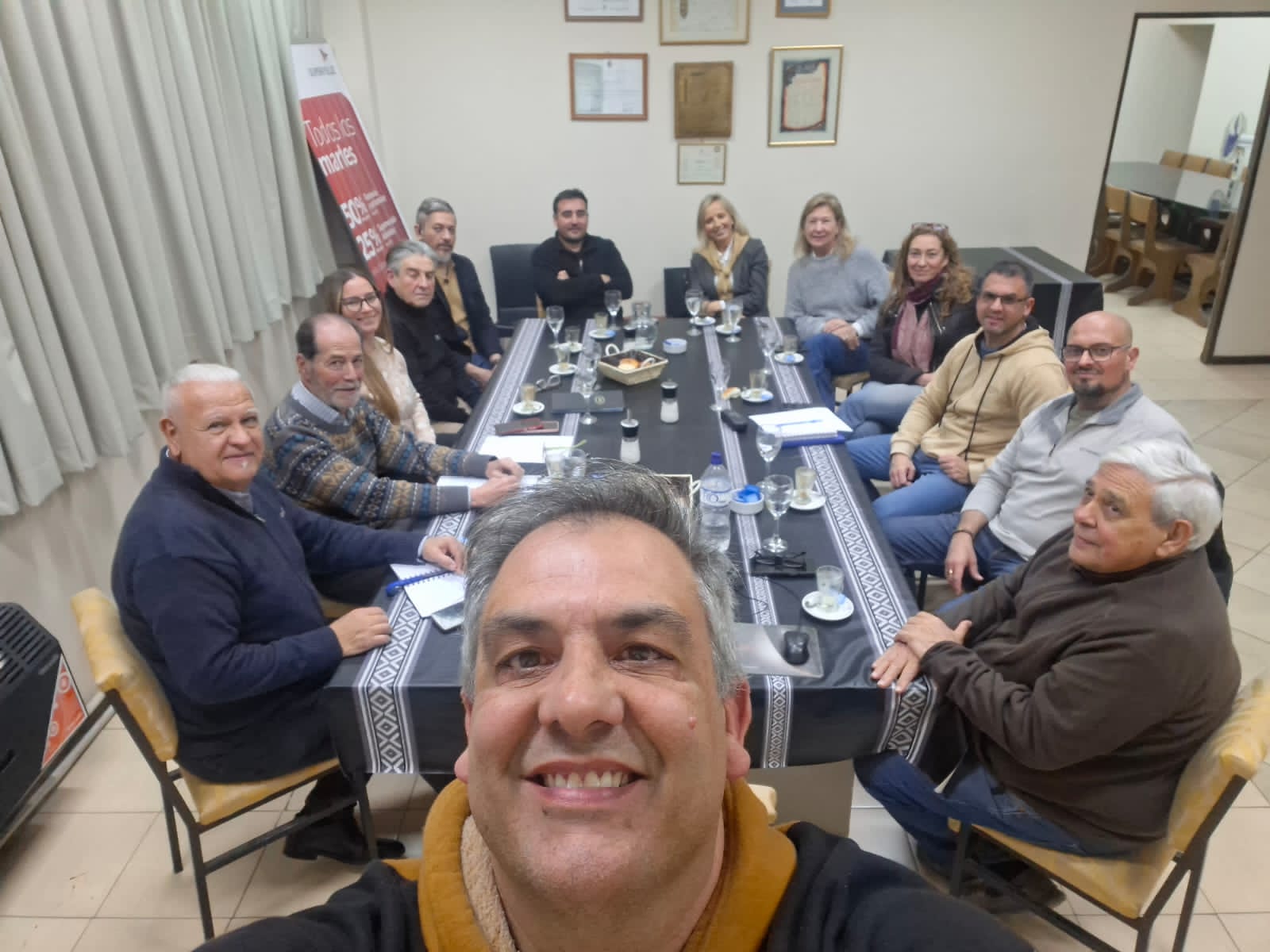 Reunión alrededor de la mesa larga, con diplomas y placas de reconocimiento en la pared